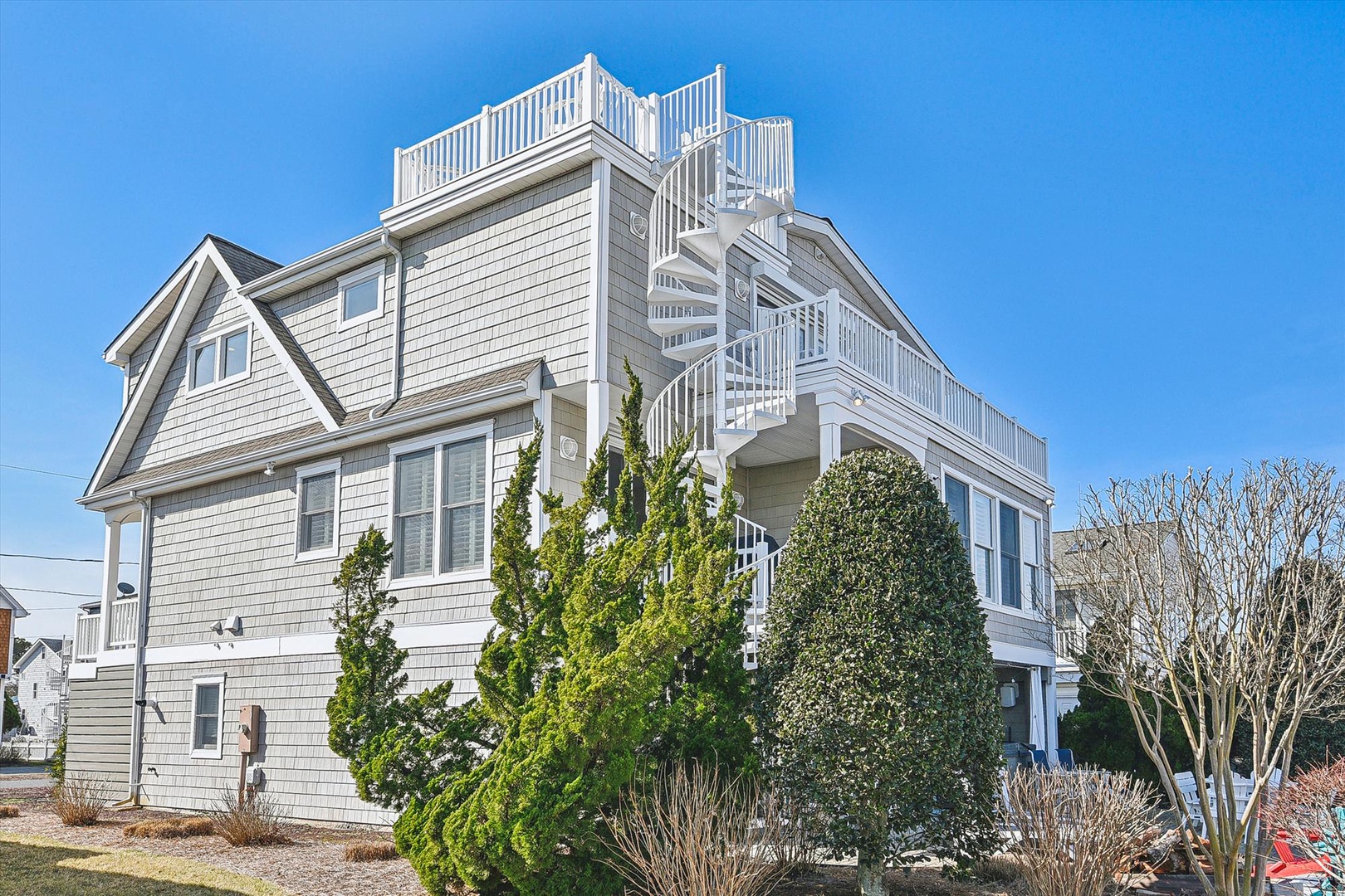 A spacious coastal property featuring white shingle siding, a rooftop deck with spiral staircase, and multiple balconies nestled among mature landscaping—ideal for seaside getaways.
