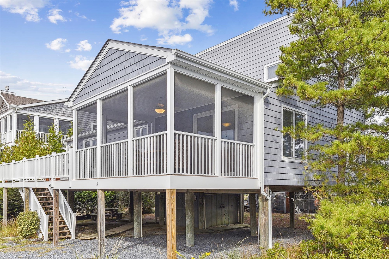 Coastal vacation home featuring gray shingle siding, elevated design with covered parking below, and a screened porch for bug-free relaxation in a beach neighborhood setting.