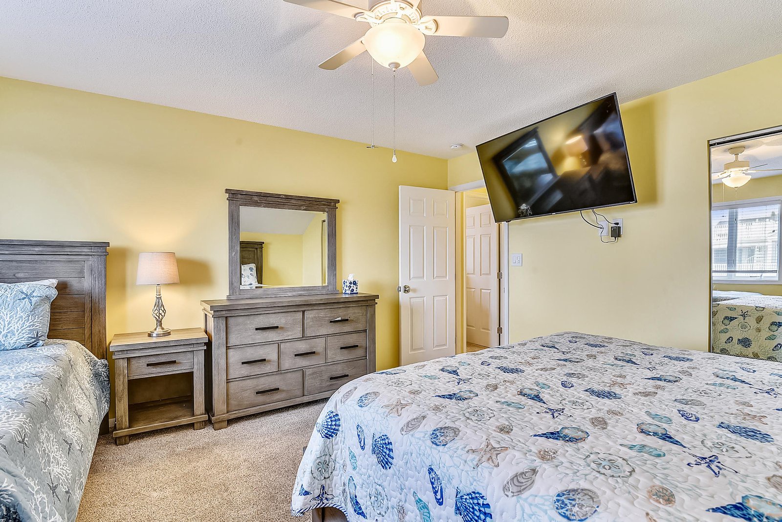 Your bright, spacious bedroom features a plush queen bed, stylish wood furniture, and a mounted TV for your entertainment. The cheerful yellow walls and ceiling fan create a warm, restful atmosphere.