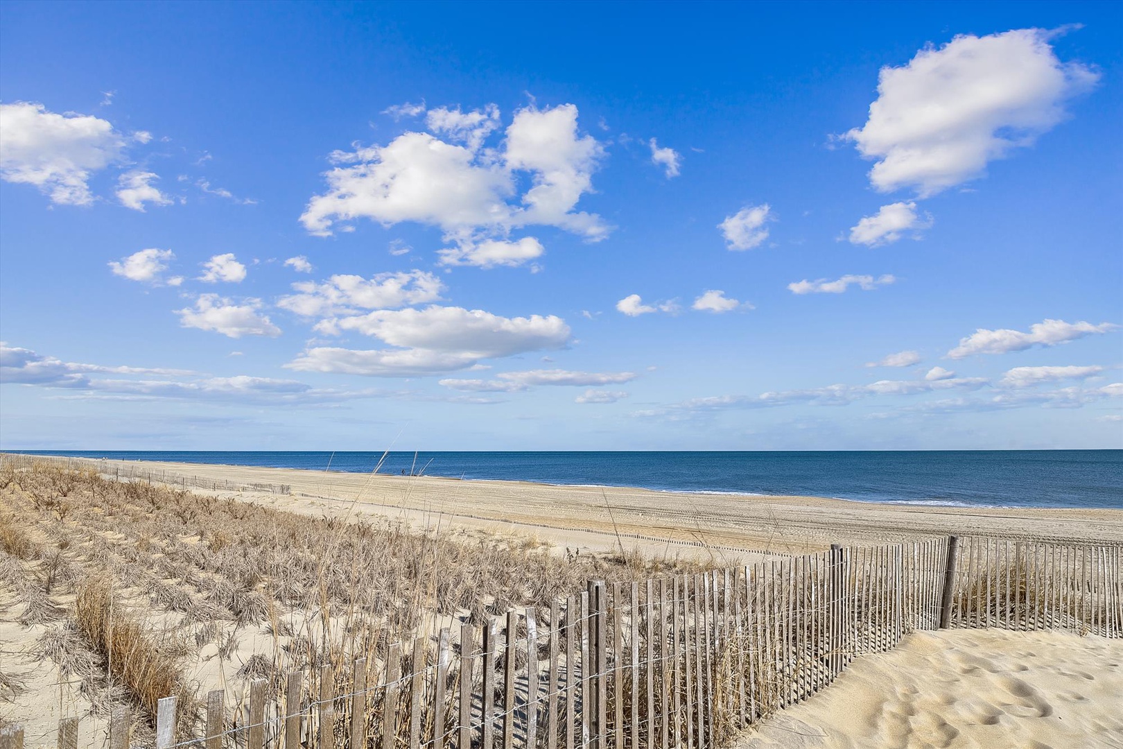A serene beachfront location with expansive ocean views, sand dunes, and weathered fencing lining the dune walkway. The property offers direct beach access and stunning coastal scenery.