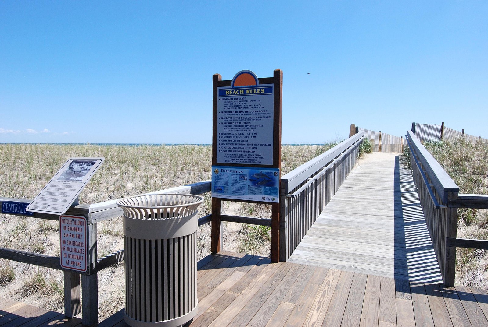 A wooden boardwalk leads through coastal dunes toward the shoreline, framed by weathered fencing and beach grass swaying in the breeze—your gateway to sun, sand, and ocean waves.