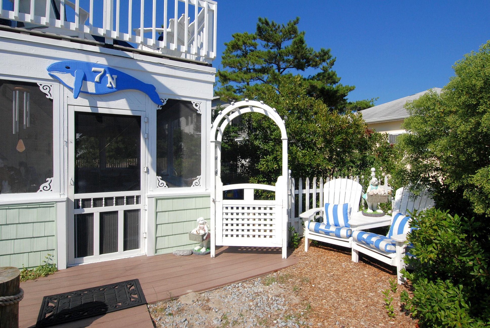 A charming coastal cottage entrance awaits, with blue-striped cushions on the outdoor seating inviting you to relax in the sea breeze beneath mature trees and a white arbor.