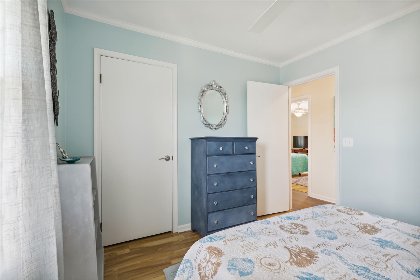 Relax in your serene coastal-inspired bedroom with soft blue walls, charming nautical touches, and a handsome dresser for your belongings. The adjacent room awaits through the open doorway.