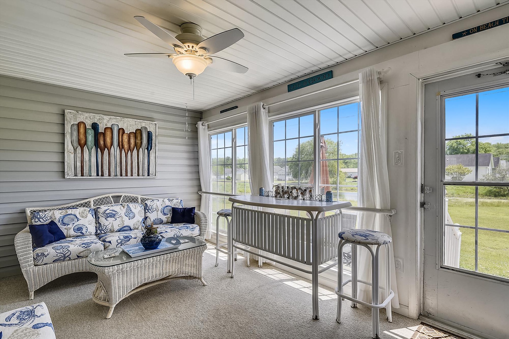 Bright coastal-themed sunroom with comfortable seating, breakfast bar, and expansive windows framing peaceful lawn and water views—perfect for morning coffee or sunset drinks.