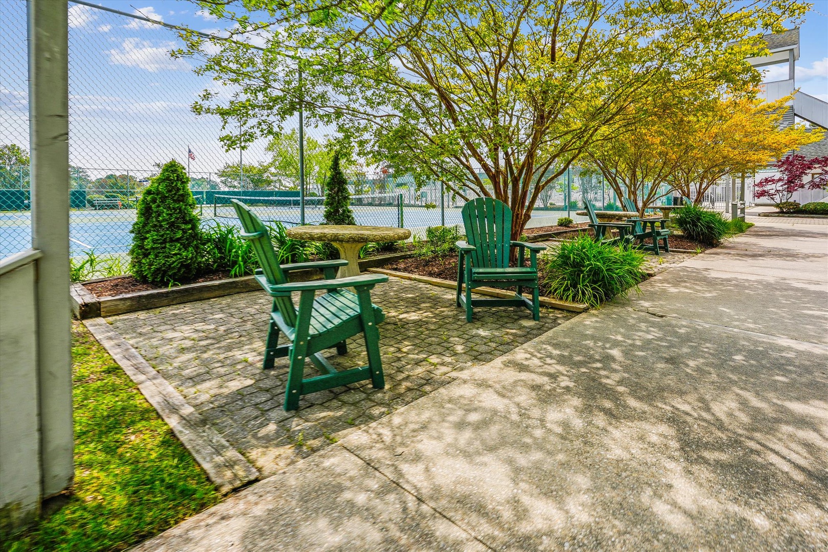 Relax in a shaded Adirondack chair overlooking the waterfront, where tennis courts and lush gardens create a peaceful outdoor retreat beneath dappled sunlight.