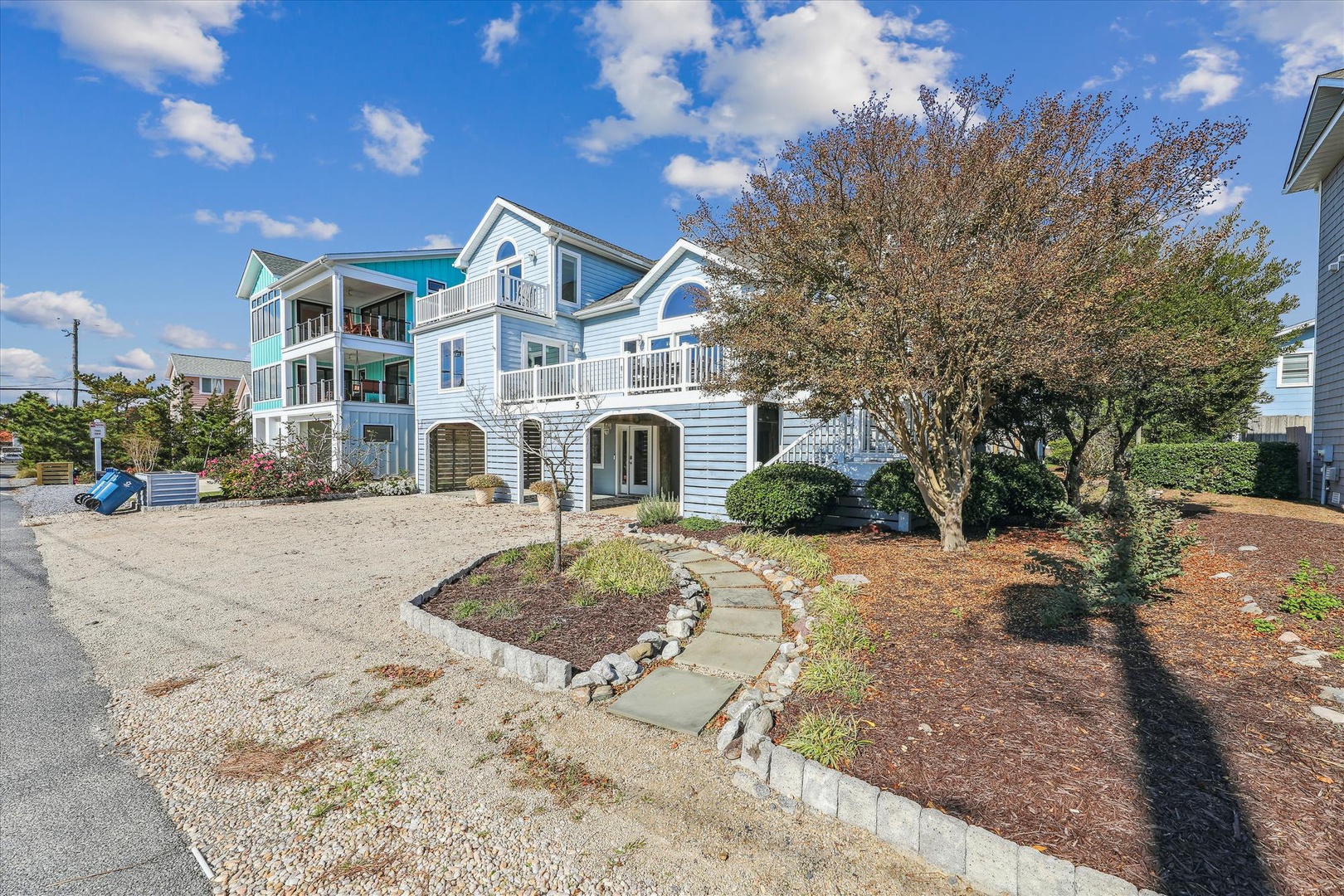 The property exterior features charming coastal architecture with multiple balconies, mature landscaping, and a welcoming stone pathway leading to your beach getaway.