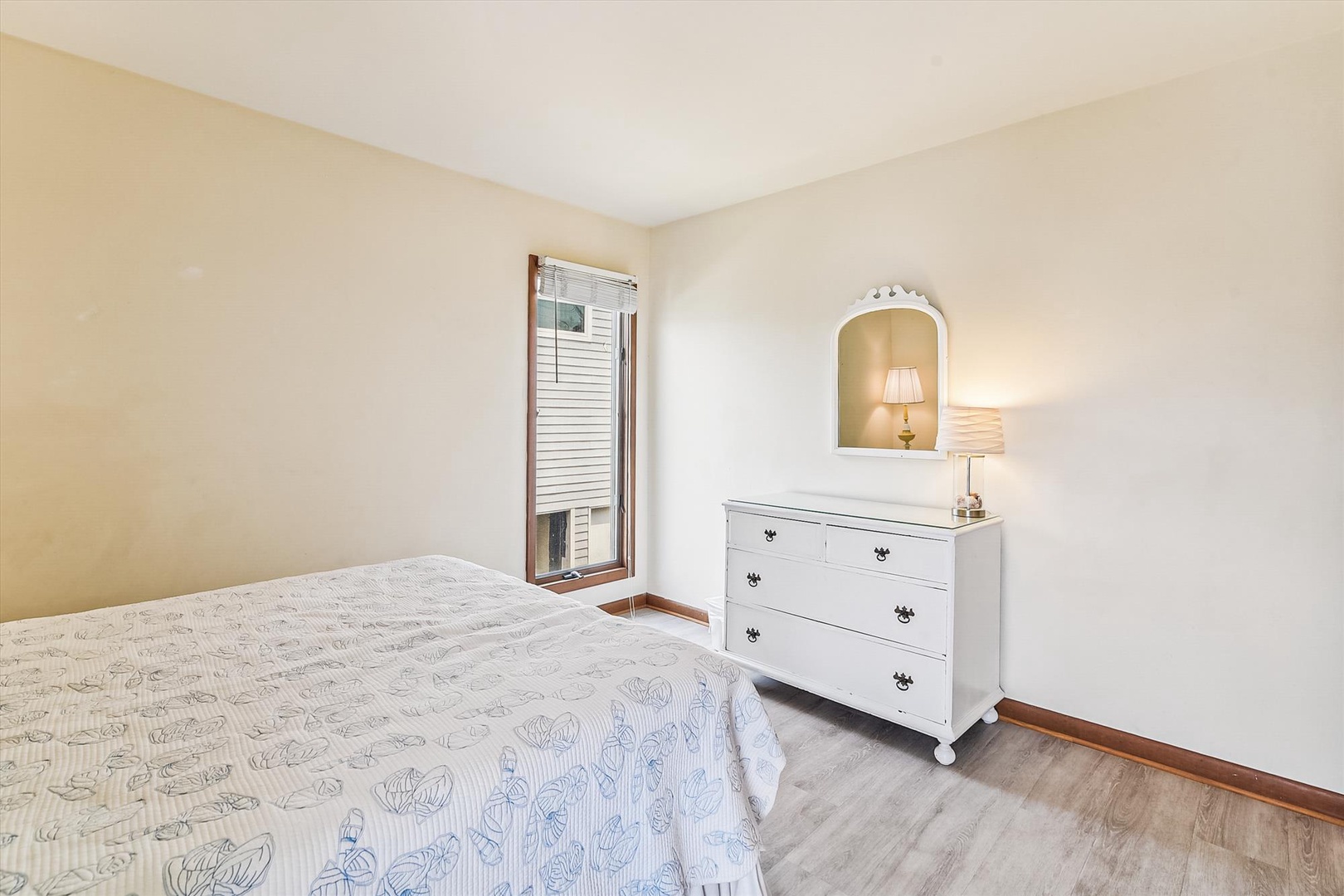 Your peaceful bedroom features soft tones, a bright window with natural light, and charming details like a vintage dresser with a warm bedside lamp.