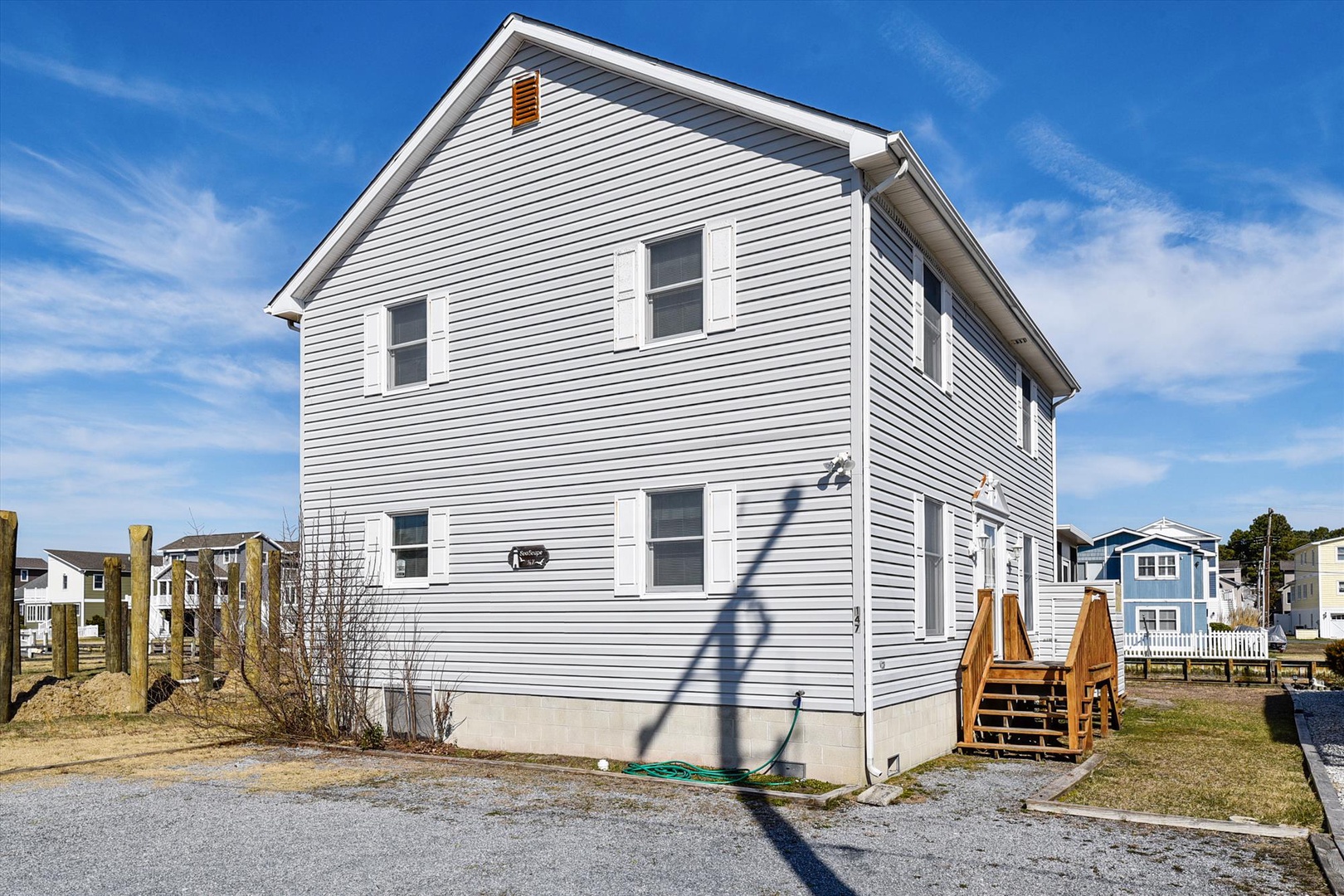 A two-story coastal vacation home with bright white siding and multiple windows, featuring wooden deck access and surrounded by neighboring beach properties under clear blue skies.