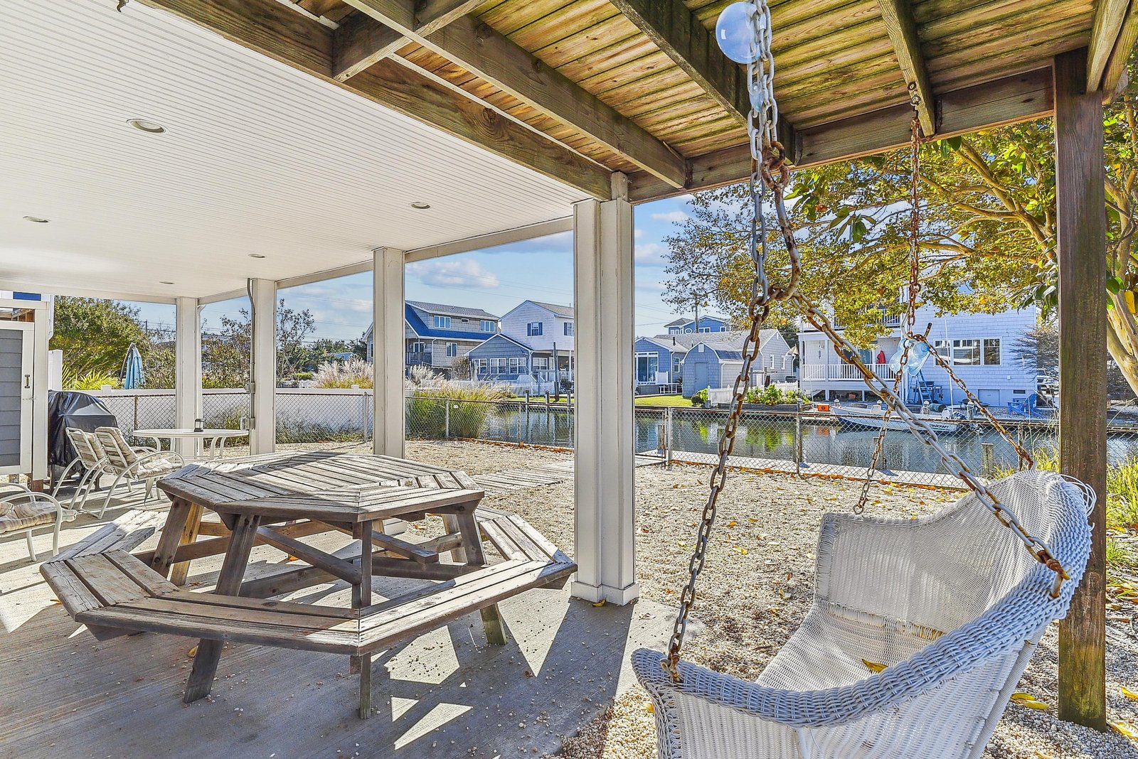 A covered patio features a picnic table and swing chair overlooking a peaceful waterfront neighborhood, perfect for relaxing after a day of coastal adventures.