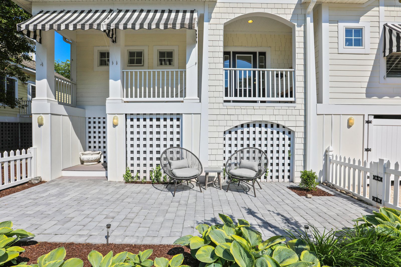 A charming coastal-style property featuring classic white shingle siding, striped awnings, and dual balconies. The brick paver patio offers a quiet spot to unwind with lattice privacy screening and cozy seating surrounded by lush greenery.