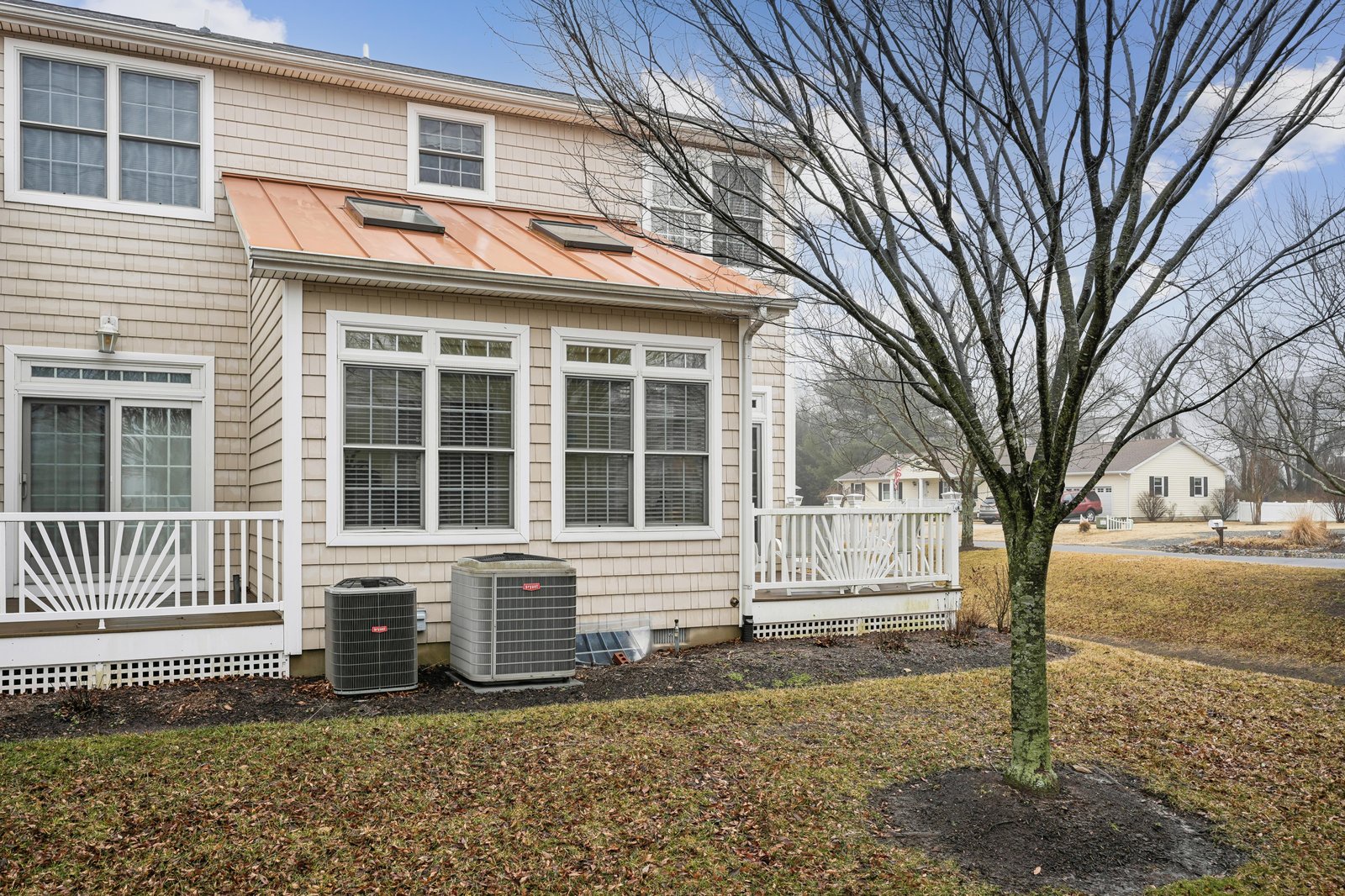 A charming beige townhome with a copper-tone metal awning, white railings, and climate control units, located in a peaceful residential neighborhood.