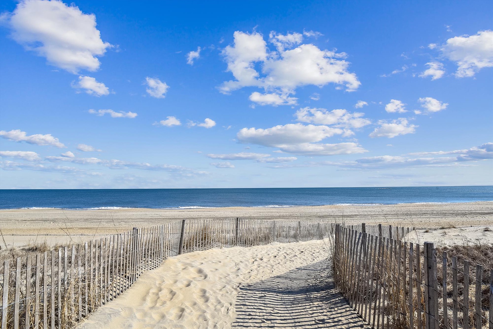 Sandy beach with weathered dune fencing stretches toward calm ocean waters beneath a bright blue sky dotted with soft clouds, offering peaceful coastal surroundings near the property.