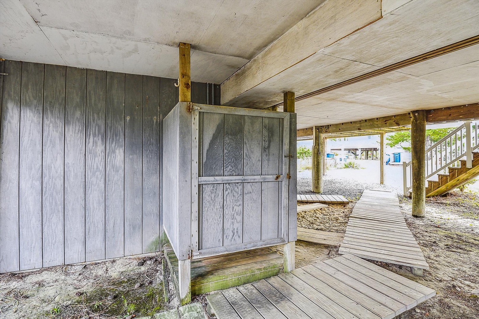 This weathered beach house sits on sturdy pilings, offering covered storage beneath the main living quarters with sandy pathways leading to nearby coastal areas.