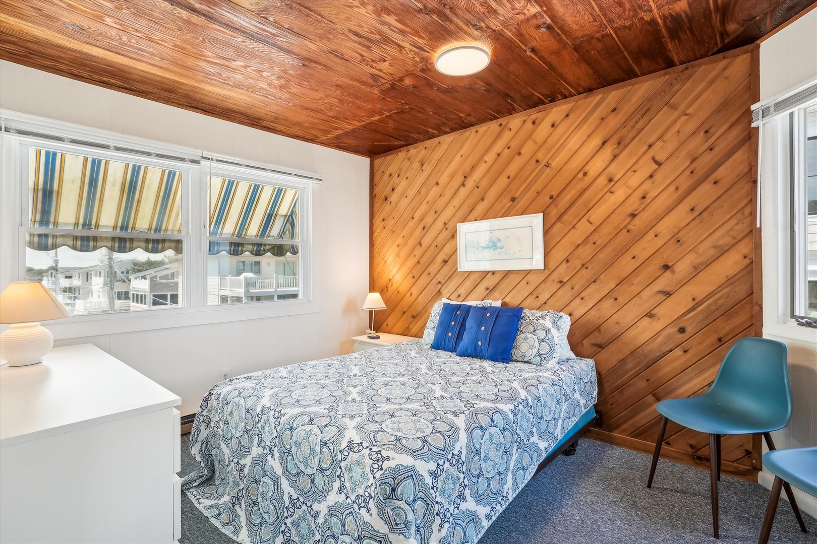 Drift off in a cozy bedroom featuring warm wood-paneled walls and a plush bed dressed in coastal blue linens. Sunlight filters through cheerful awnings while you unwind in this charming retreat.