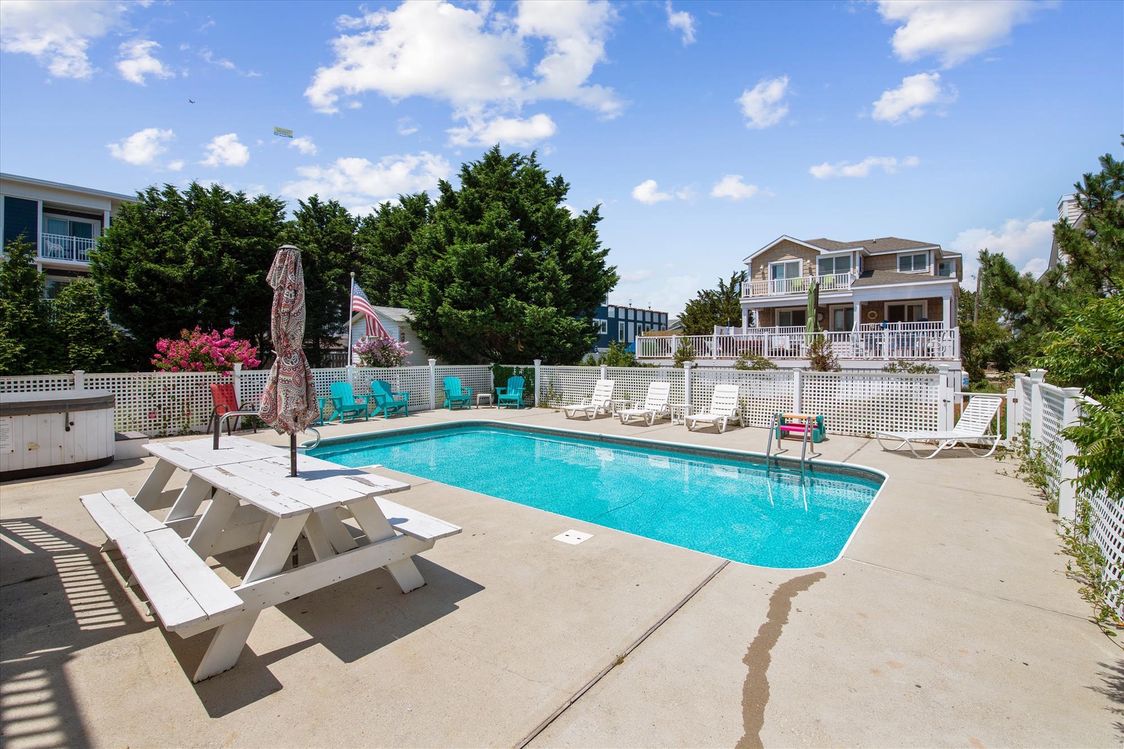 The sparkling pool area features vibrant turquoise and white lounge chairs, surrounded by lush greenery and charming neighborhood views under a bright blue sky.