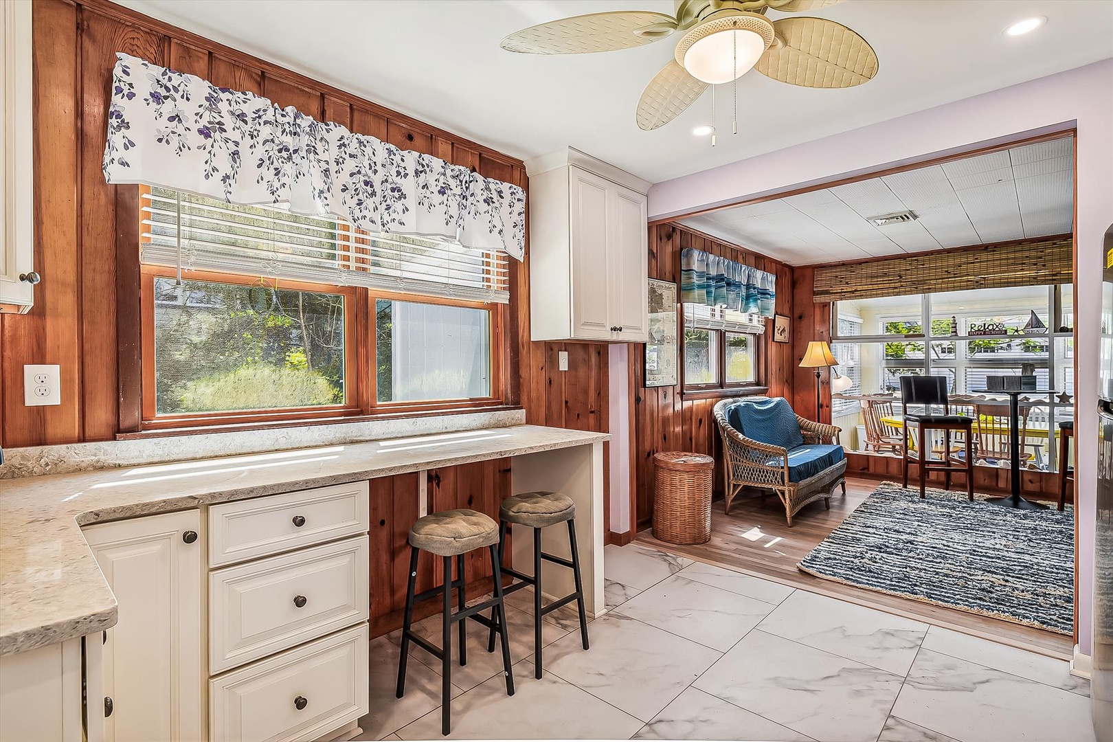 Step into your coastal retreat with warm wood paneling, a breezy ceiling fan, and charming wicker seating. Natural light fills the space, inviting you to relax and feel at home.