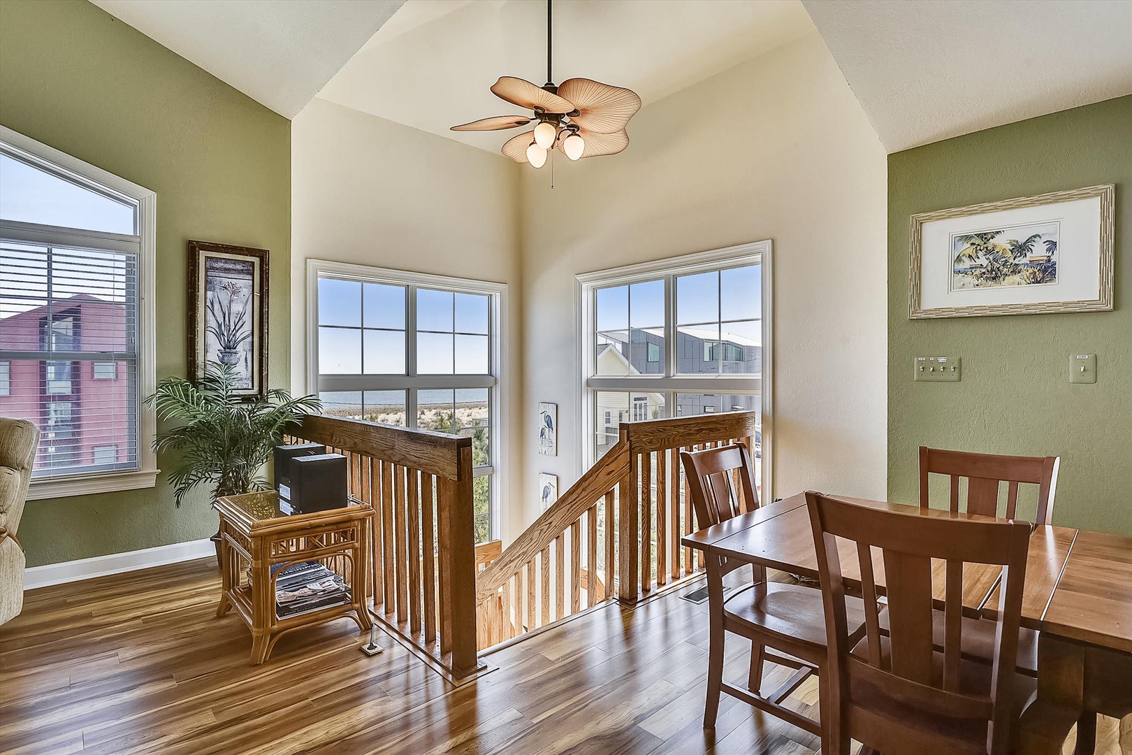 This bright, vaulted space combines seating and dining areas with warm wood floors and tropical-style ceiling fan. Large windows invite natural light and ocean glimpses beyond the neighboring homes.