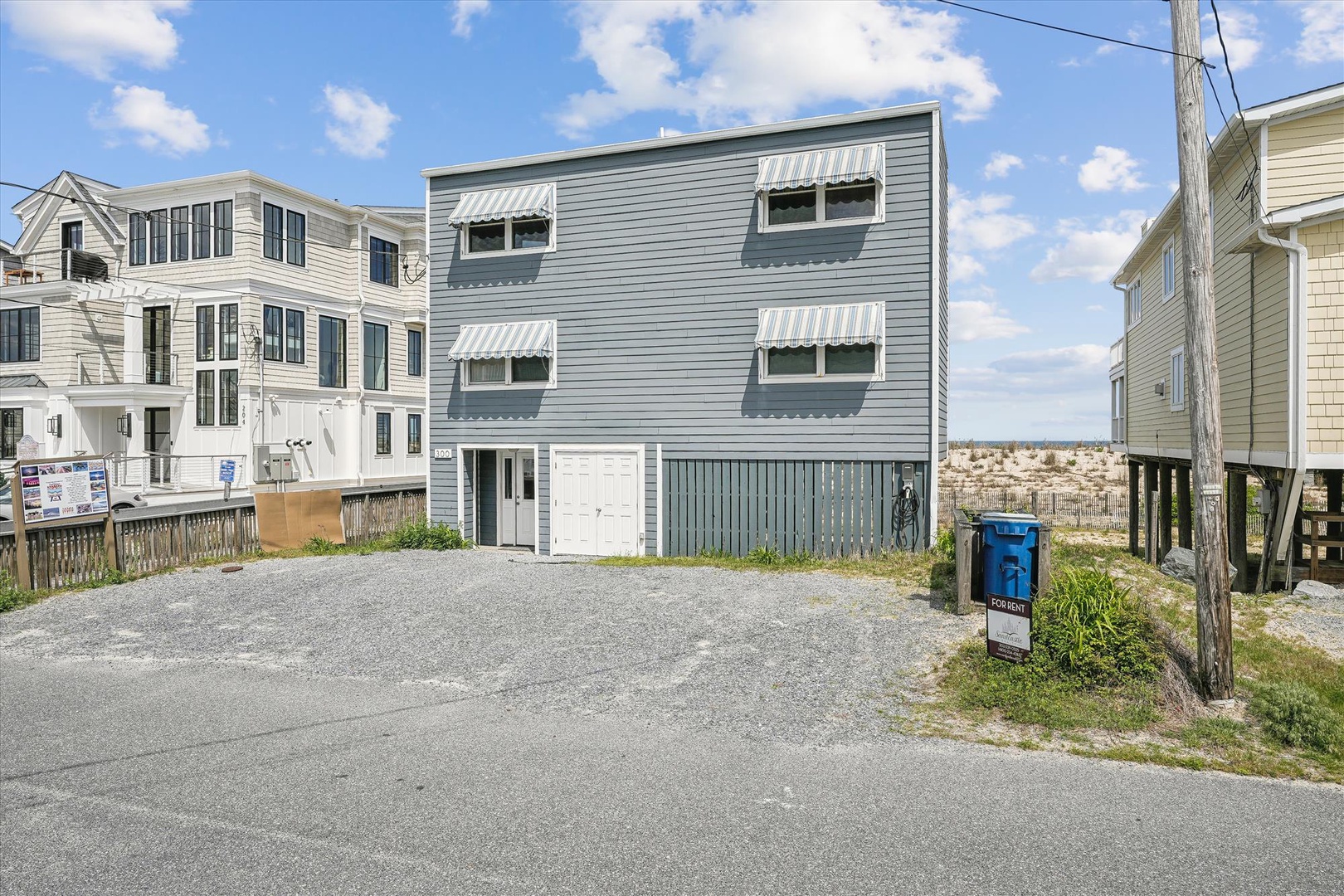 Gray beach house elevated on stilts, located in a coastal neighborhood with nearby beach access. Property features dedicated parking and striped window awnings.