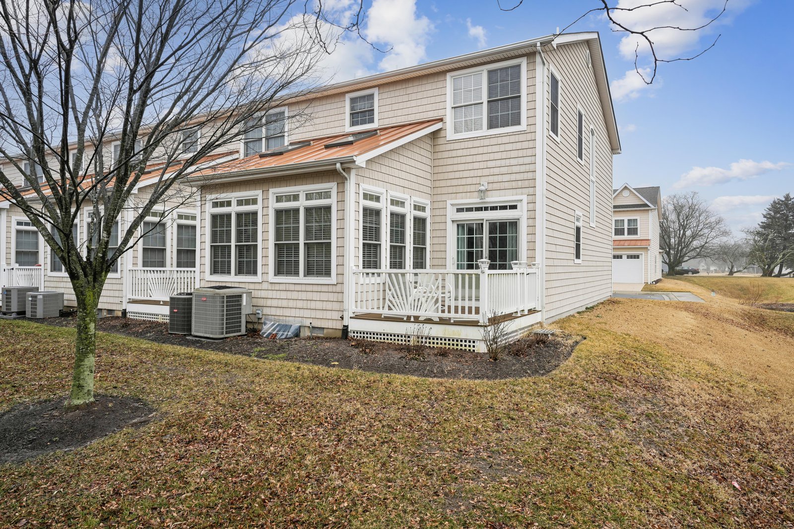 A charming two-story coastal-style property featuring classic neutral siding, a spacious sunroom with abundant windows, and a welcoming porch entrance overlooking peaceful seasonal grounds.