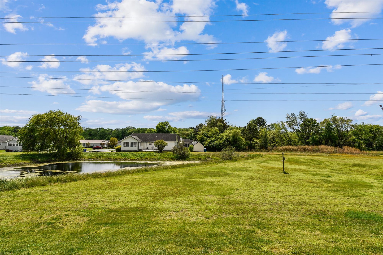 A peaceful rural setting features open green lawns, a small pond, and neighboring homes nestled among mature trees under a blue sky—the property's quiet countryside location.