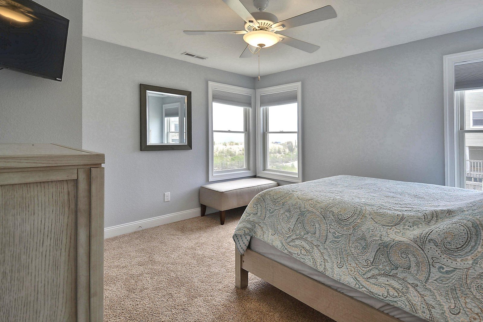 Your serene bedroom retreat features soft gray walls, a ceiling fan for comfort, and inviting natural light from corner windows that frame peaceful neighborhood views.