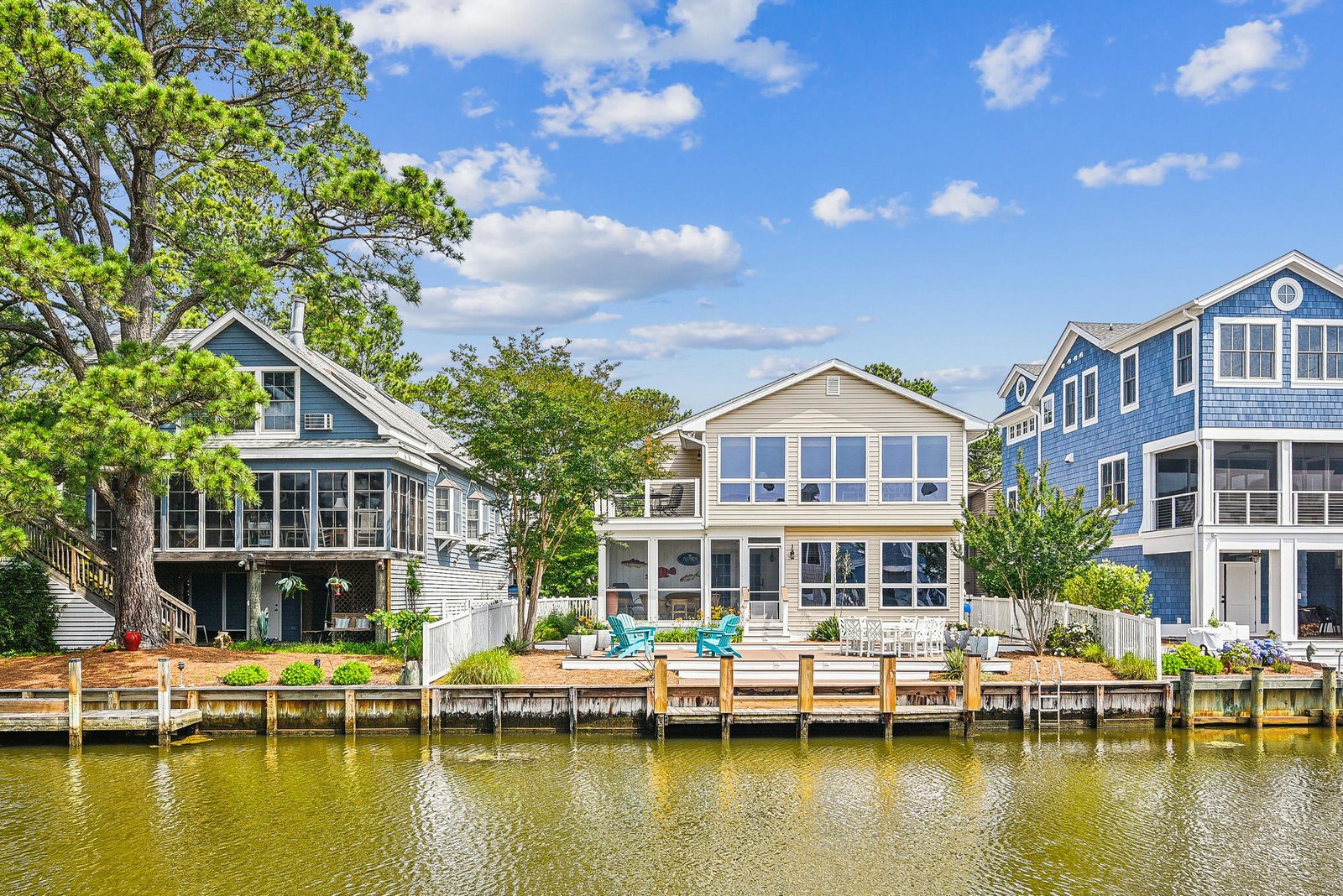 Waterfront coastal homes featuring private docks on a calm canal, surrounded by mature trees and lush landscaping under a bright blue sky.