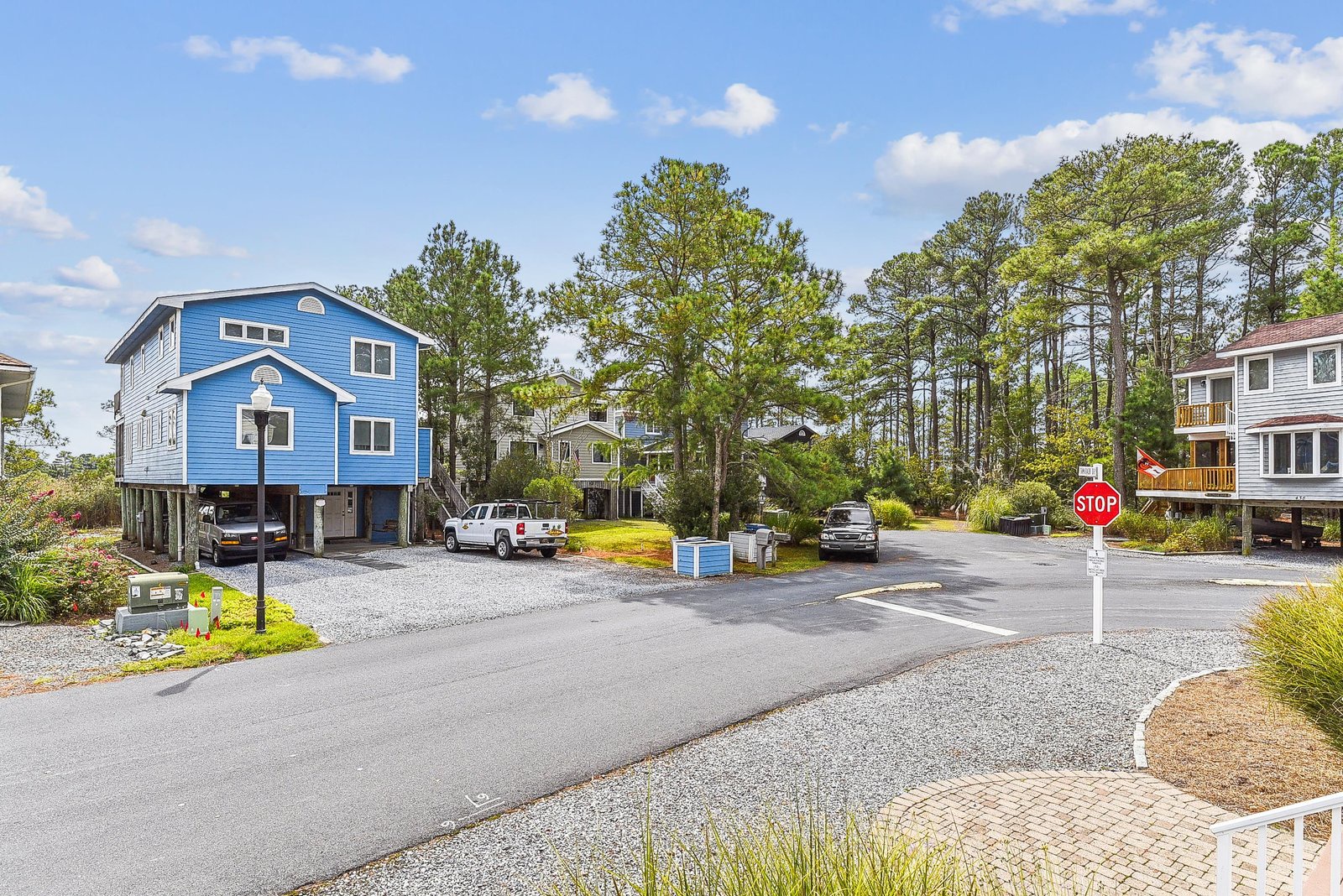 A coastal neighborhood street showcasing the property—a distinctive blue beach house with parking and lush green surroundings.