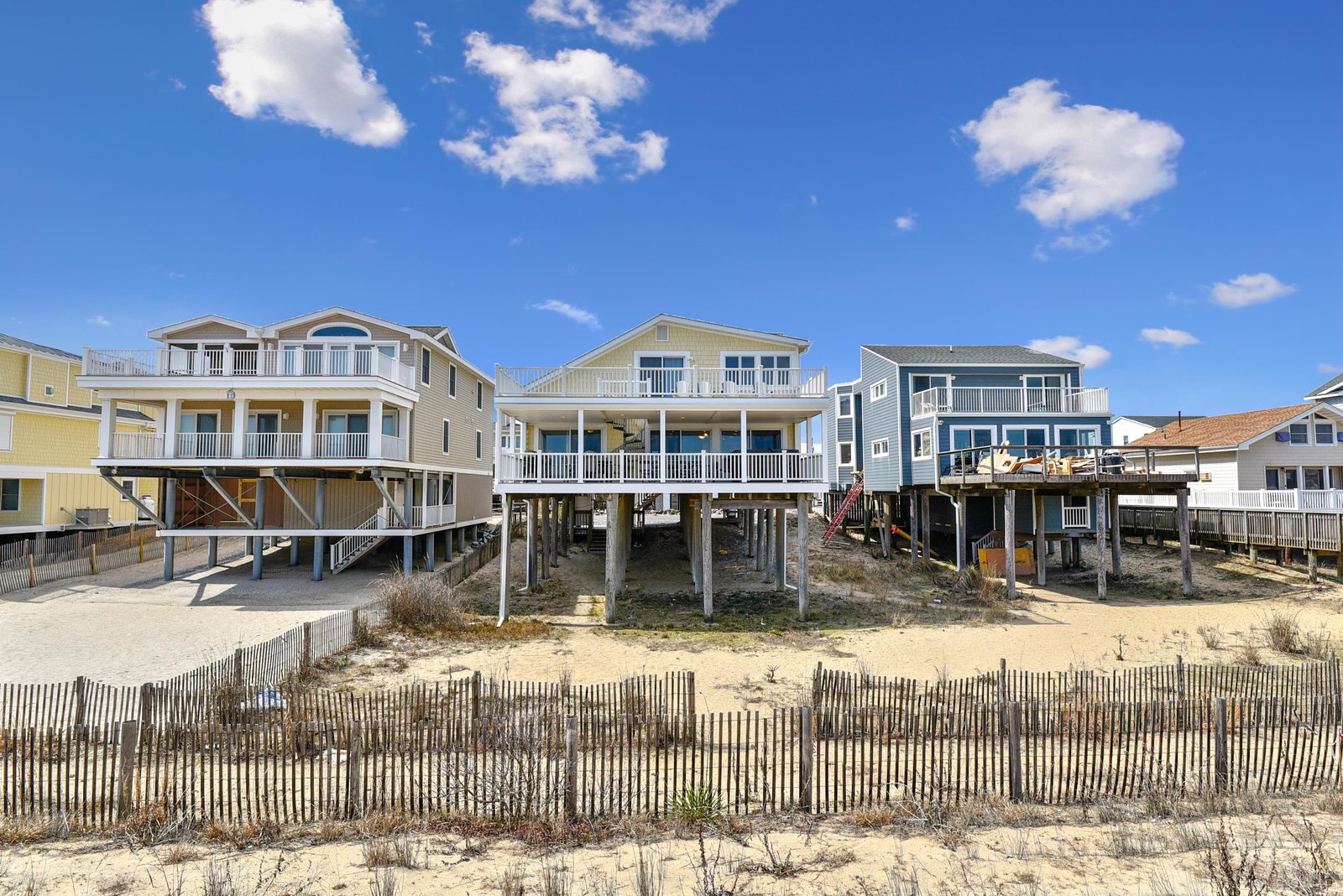 Elevated beachfront homes with direct beach access, situated along a sandy shoreline with protective dune fencing and clear blue skies overhead.
