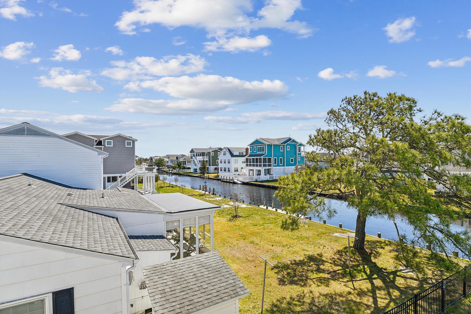 Charming waterfront neighborhood featuring canal-front homes, lush green spaces, and mature trees under a bright blue sky with scattered clouds.
