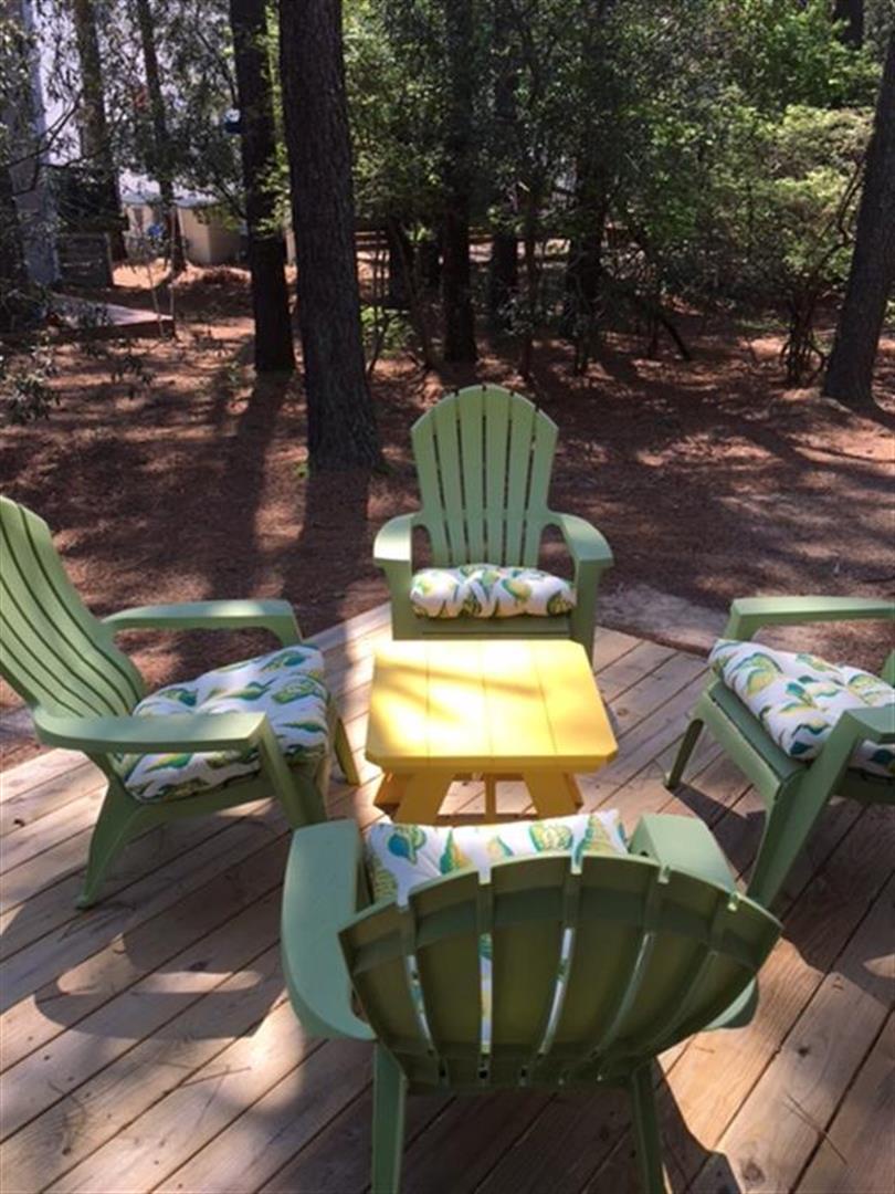 Settle into the shade of towering pines on your private deck, where bright Adirondack chairs and a cheerful table invite lazy afternoons surrounded by peaceful woodland.