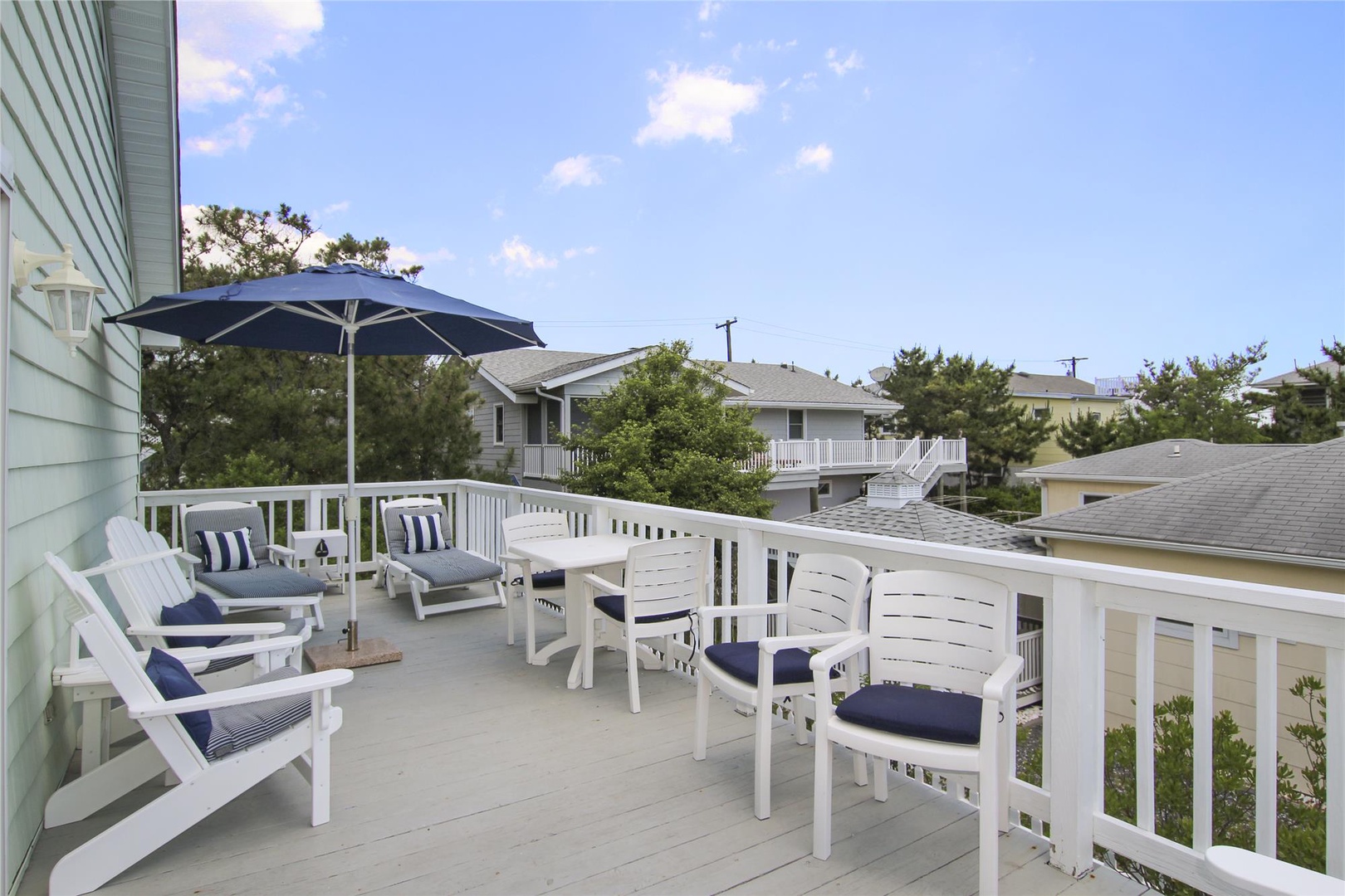 Spacious deck features multiple Adirondack chairs with navy cushions and a shaded umbrella, perfect for morning coffee or evening relaxation in the coastal air.