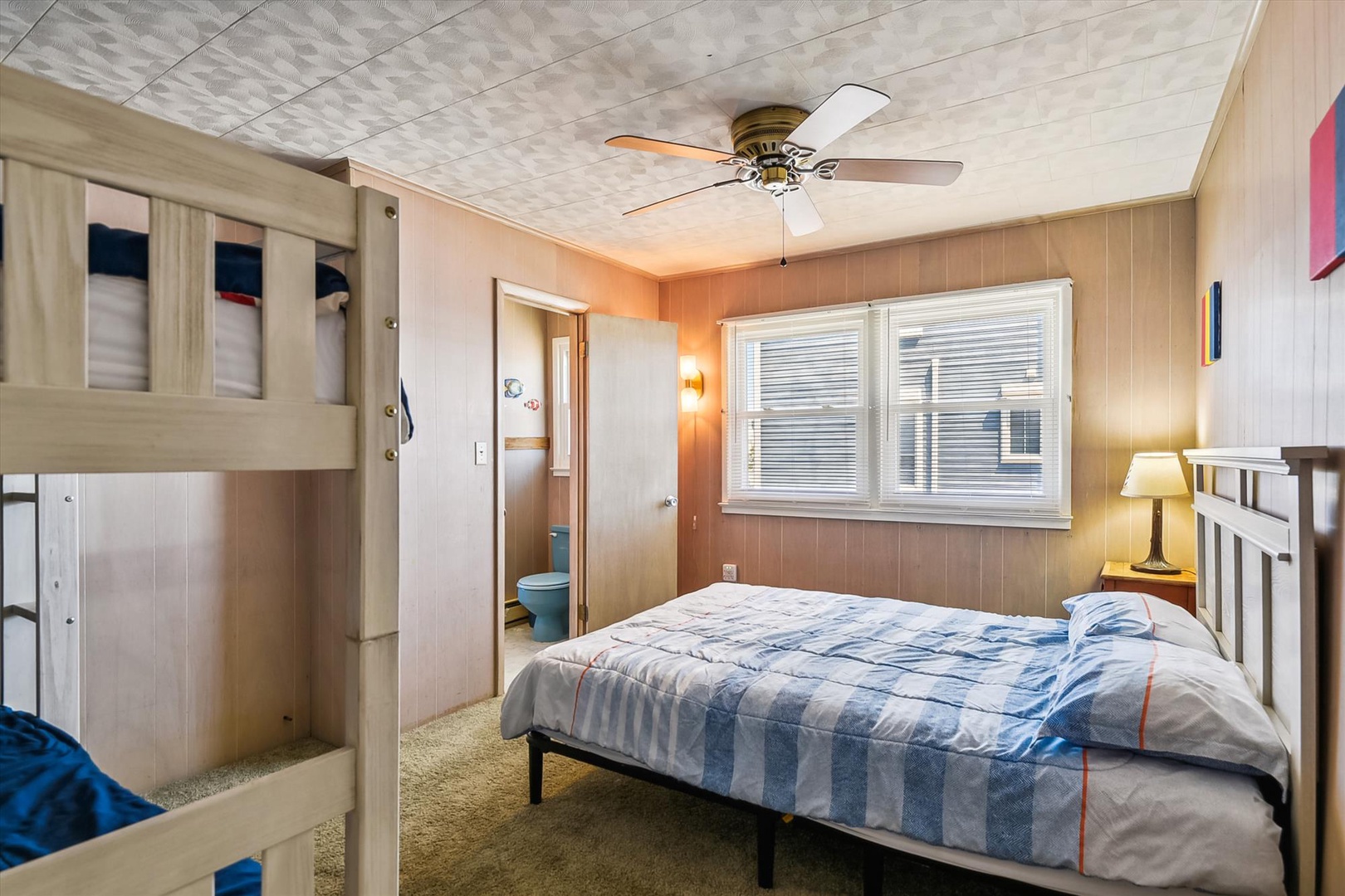 Unwind in your cozy bedroom with warm wooden walls, a ceiling fan to keep you cool, and gentle natural light streaming through the windows for a peaceful night's sleep.