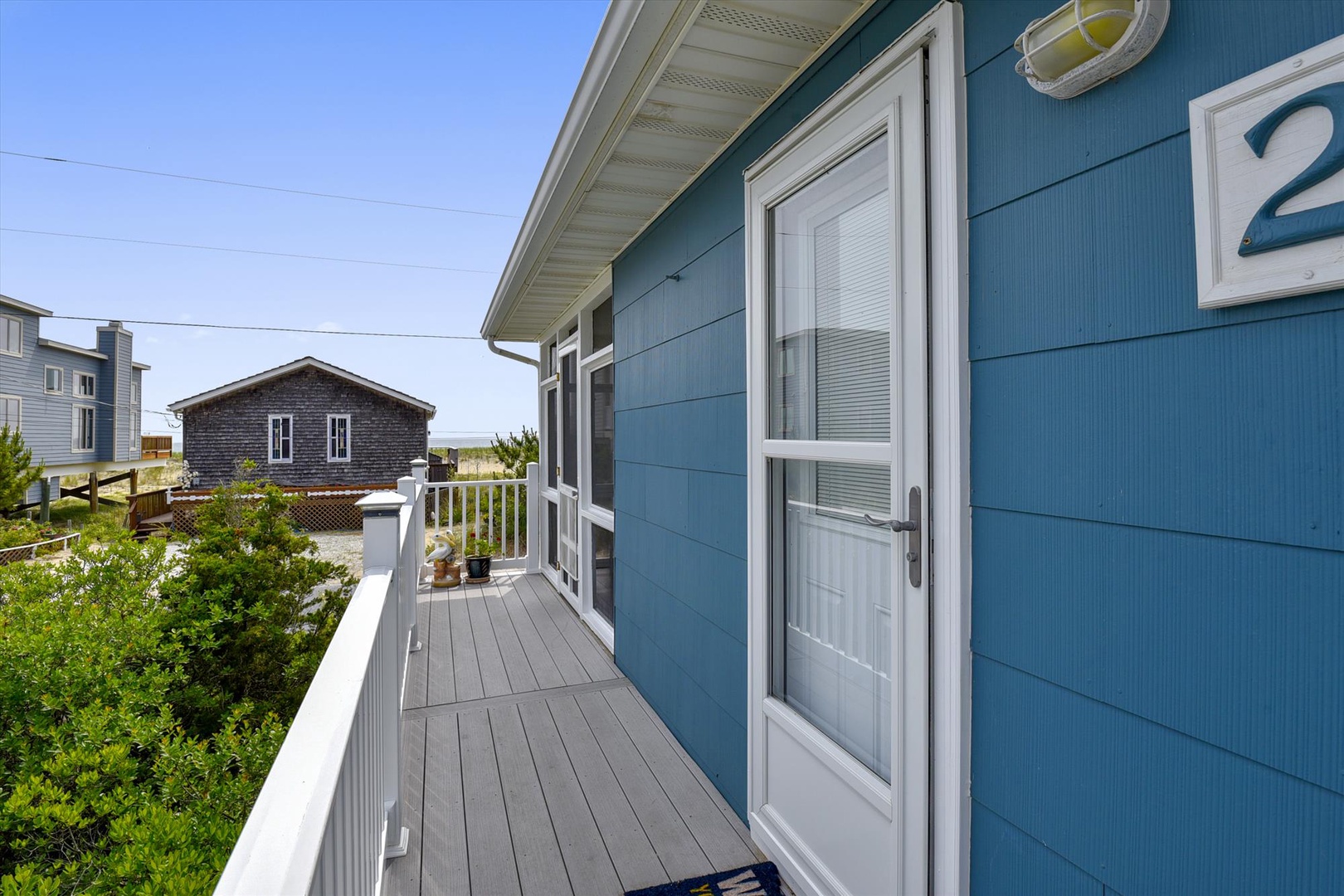 Charming blue coastal cottage exterior featuring a covered side balcony with white railings and neighboring beach homes visible in the seaside community backdrop.