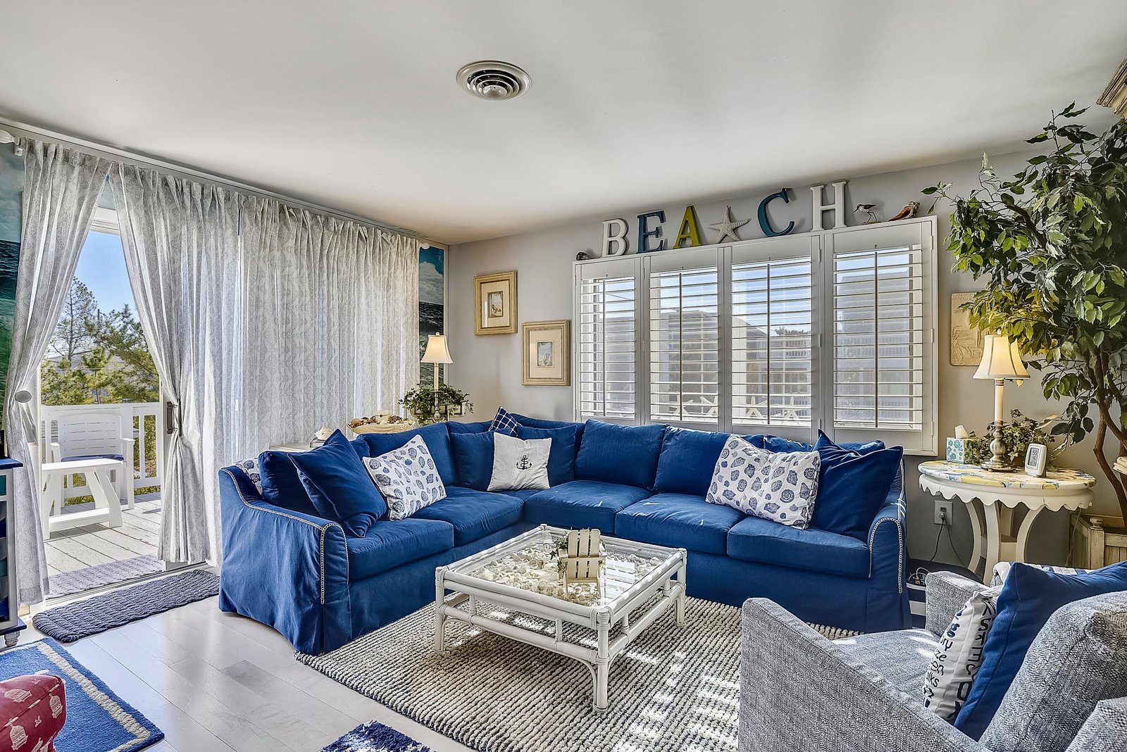 Sink into soft seating as natural light pours through white shutters and the open balcony door. Beach-themed accents and cheerful blue tones create a bright, welcoming retreat.