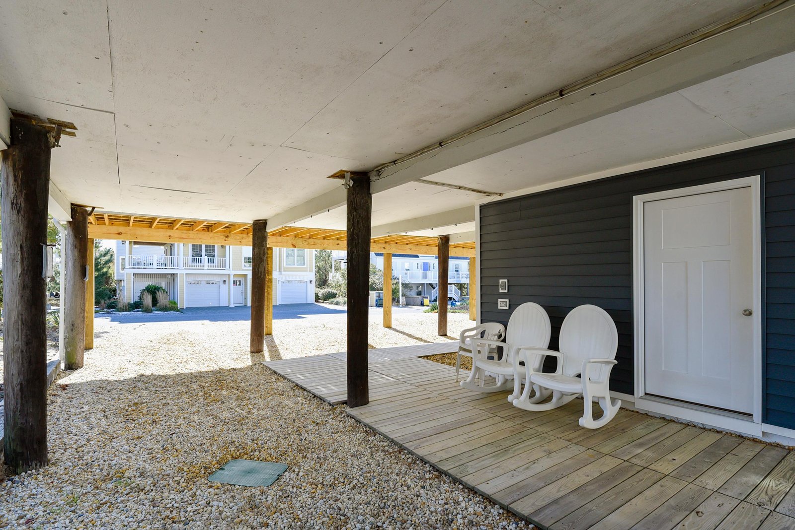 Covered ground-level area with wooden decking and casual seating, providing shaded outdoor space beneath the elevated structure. Gravel surrounds and street-facing views connect you to the neighborhood.