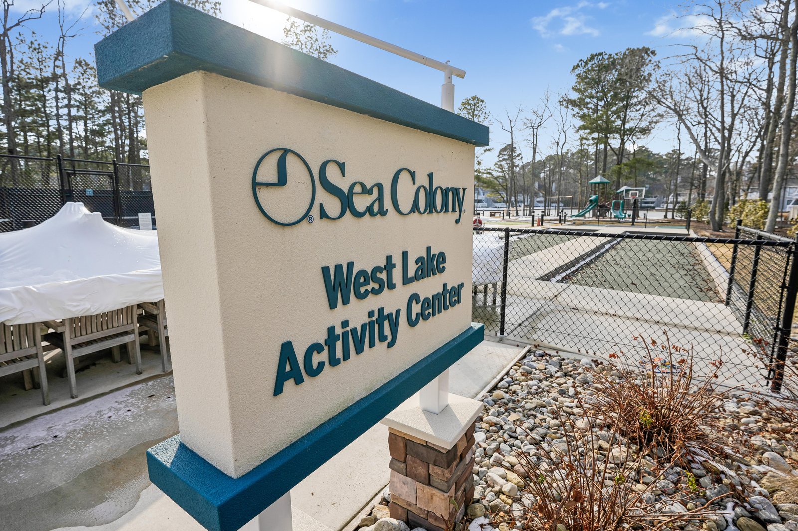 The property entrance marker stands beside tennis courts and a playground within the Sea Colony resort community, surrounded by mature pine trees.