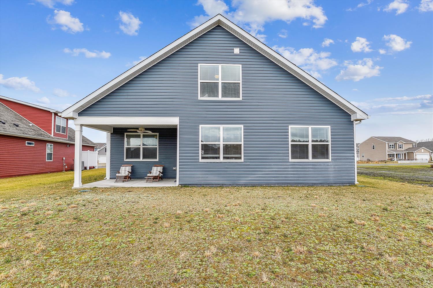 Spacious blue siding home with a covered porch featuring two lounge chairs, perfect for relaxing. Set on a large lawn in a peaceful neighborhood with nearby properties visible.