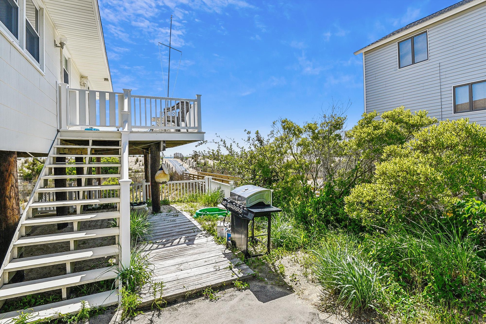 Multi-level deck leads through lush coastal greenery to ocean access, complete with BBQ grill for beachside cookouts under sunny skies.