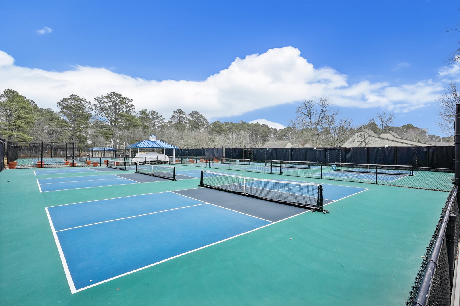 Multiple tennis courts with professional nets and markings, surrounded by mature trees under bright blue skies.