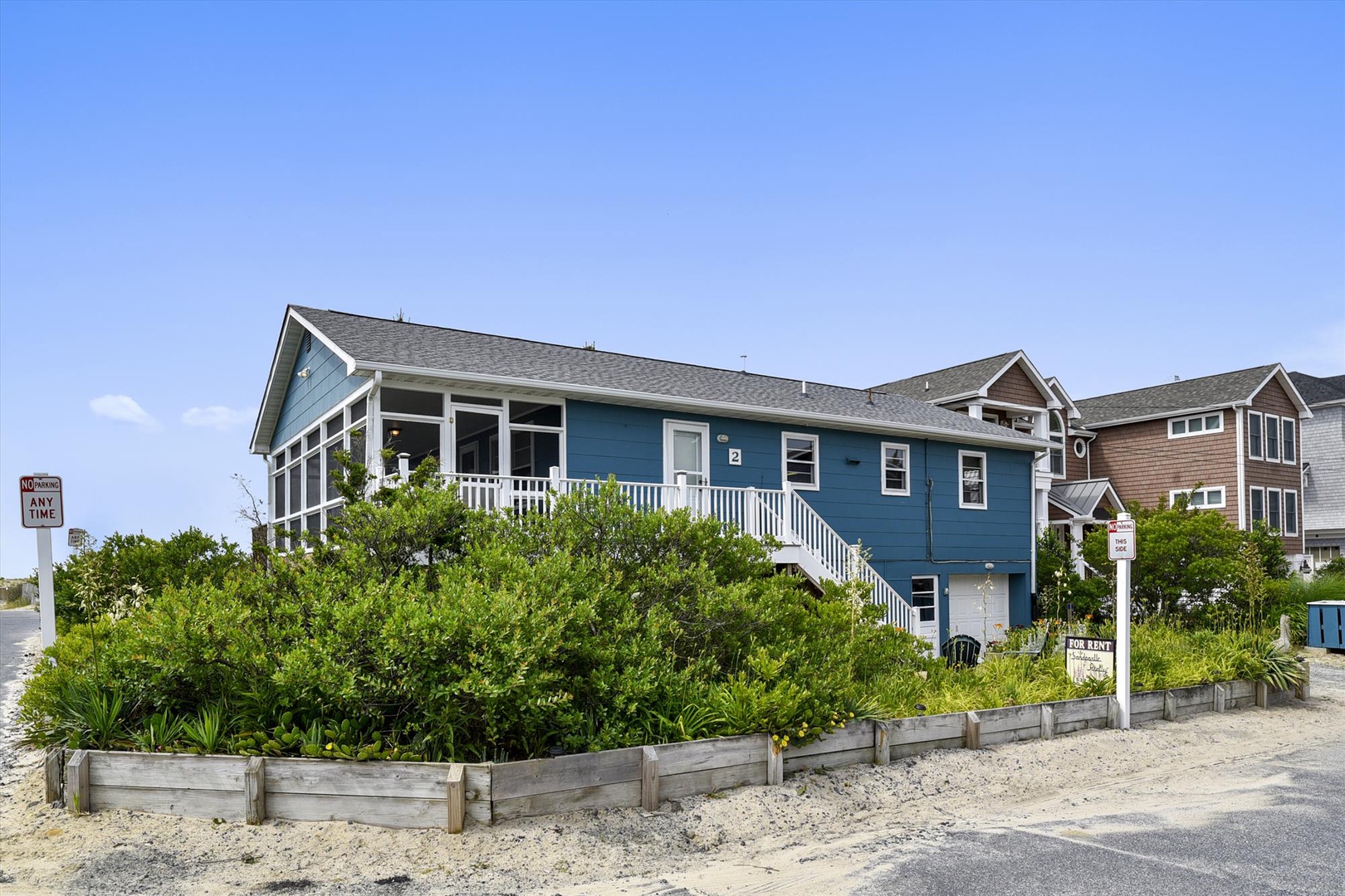 The charming blue beach house with white trim sits steps from the sand, offering coastal living in a peaceful neighborhood. Lush greenery frames the property's welcoming entrance.