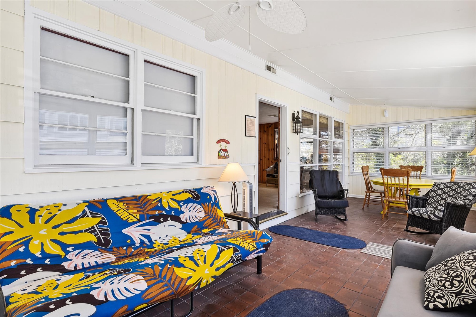Step into your bright, cheerful living area where a vibrant tropical daybed invites you to unwind. Sunlight pours through large windows, illuminating the cozy seating and warm terracotta floors that connect you to the dining space beyond.