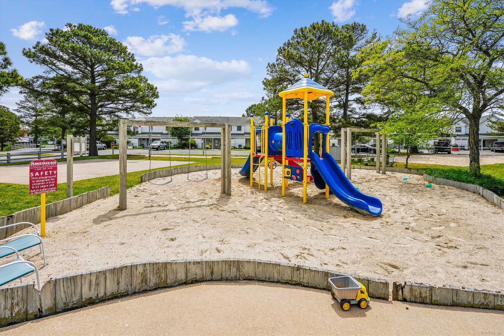 A bright playground with slides and climbing structures sits in a sandy play area, surrounded by mature trees and residential buildings, offering families a welcoming outdoor space.