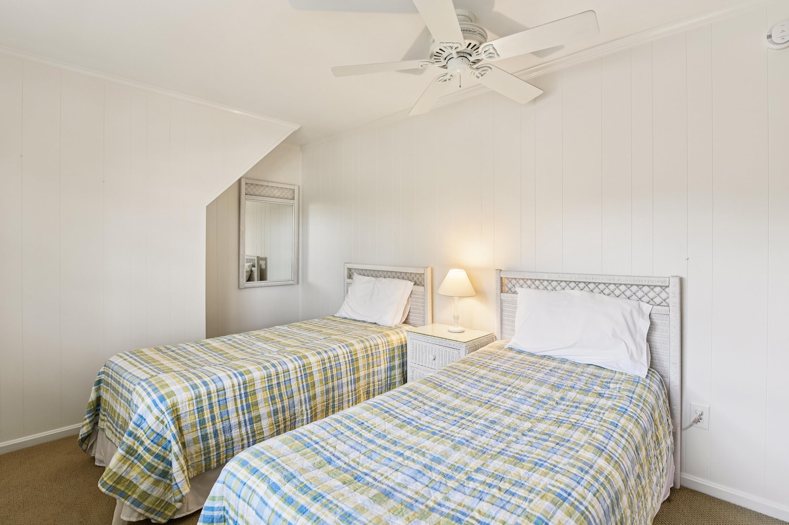 Rest easy in your cozy twin bedroom with crisp linens and gentle ceiling fan. White walls and soft plaid blankets create a peaceful space for your vacation dreams.