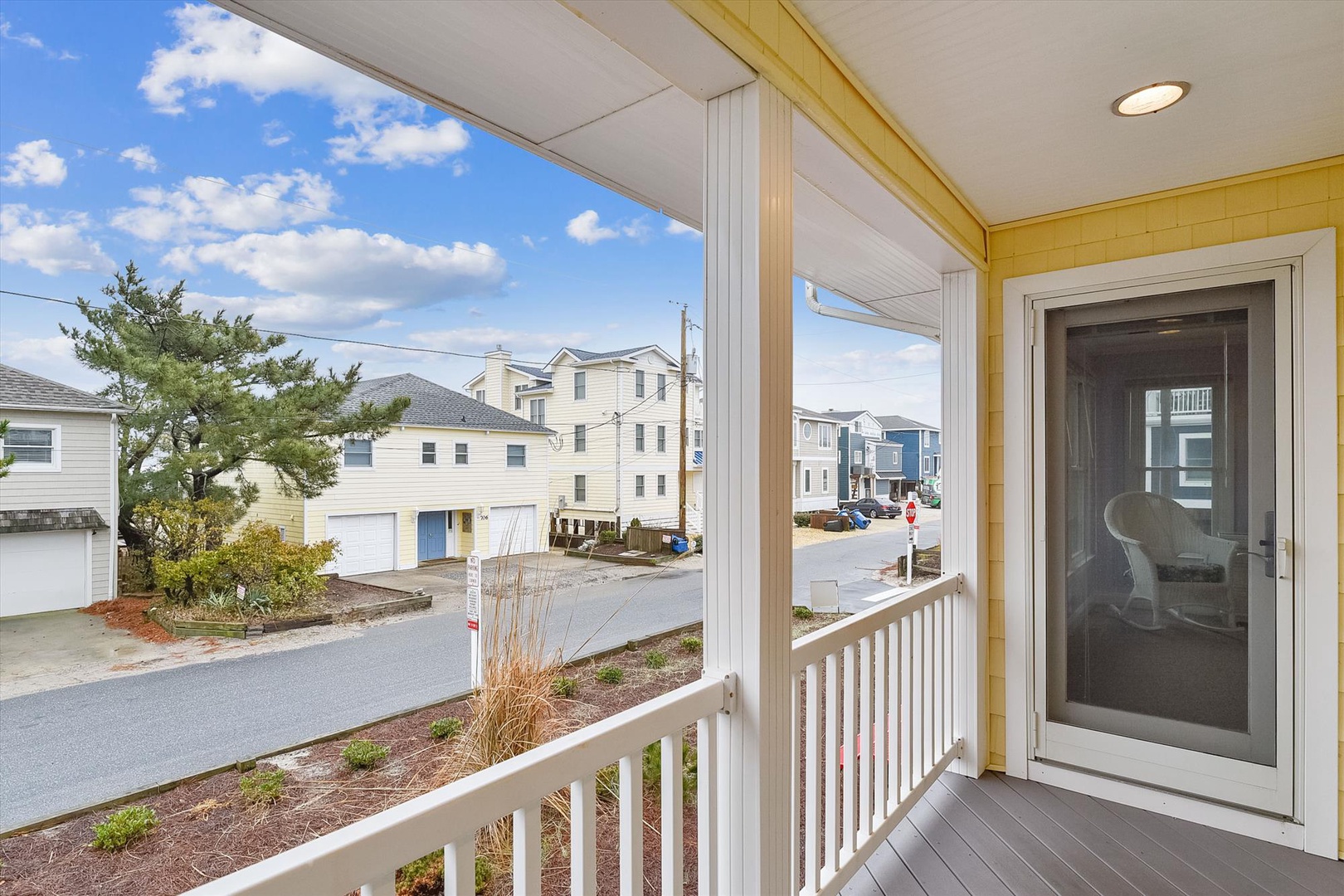 A covered porch with white railings overlooks a peaceful coastal neighborhood lined with beach houses beneath a bright blue sky.