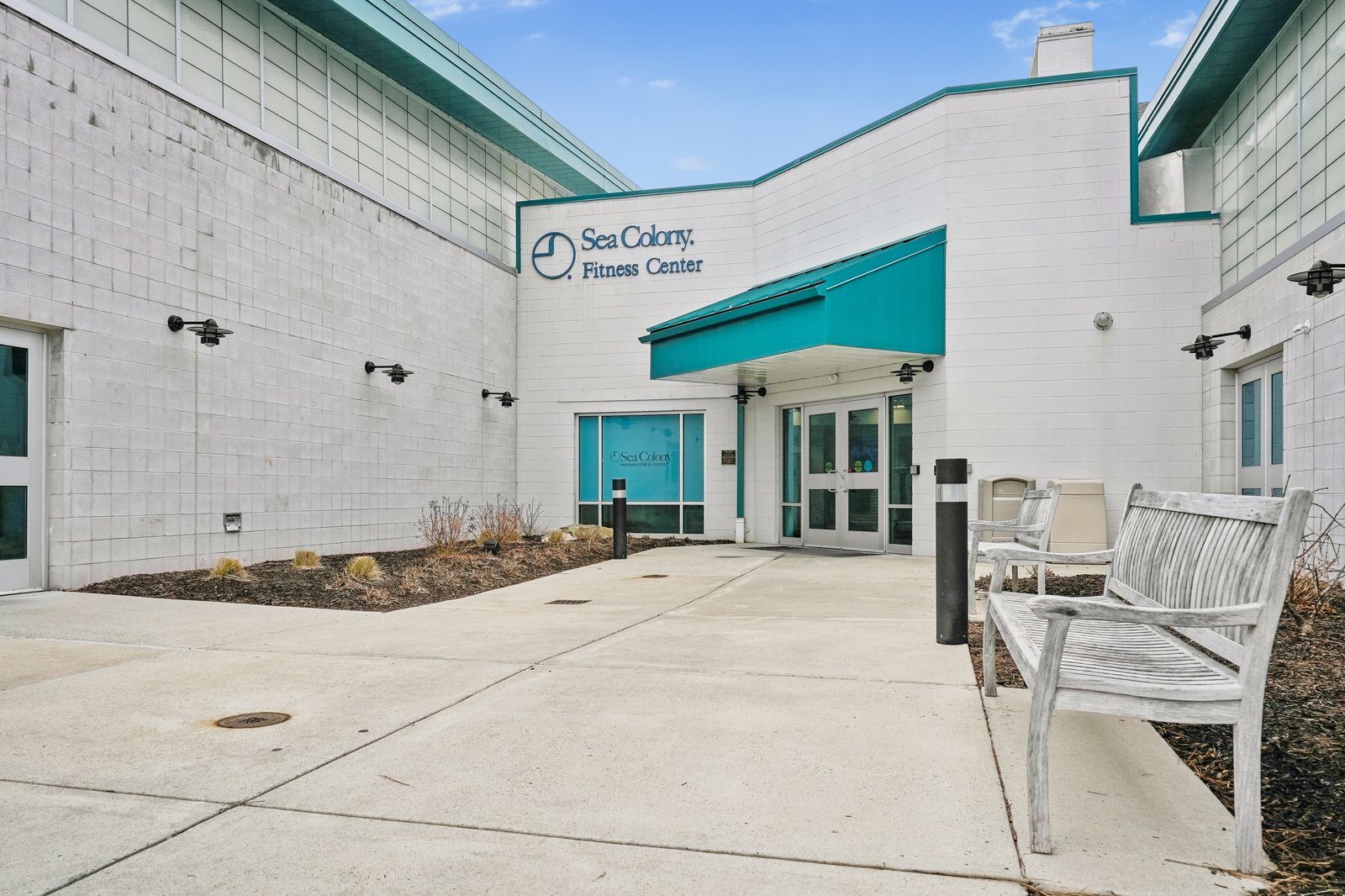 Sea Colony Fitness Center entrance with teal accents and outdoor seating area under blue skies.