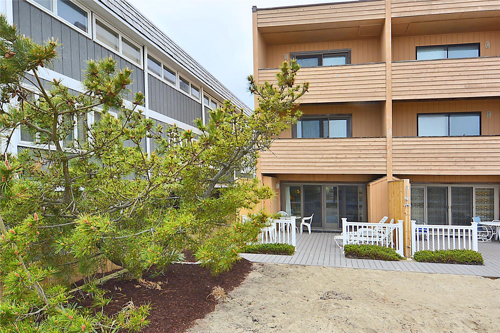 Modern wood-sided property exterior with ground-level units featuring private patios and white outdoor furniture, framed by coastal landscaping and mature trees.