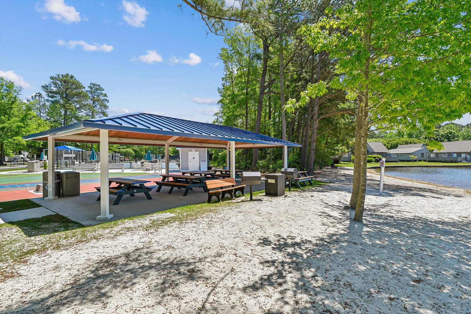 Covered pavilion with picnic tables and BBQ facilities sits beside a sandy lake beach area surrounded by mature trees.