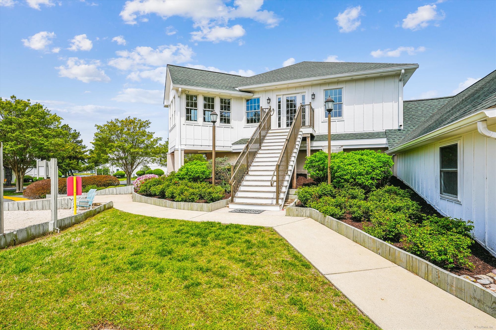 A white coastal-style property with exterior staircase and well-maintained gardens set in a peaceful location with water views nearby under blue summer skies.