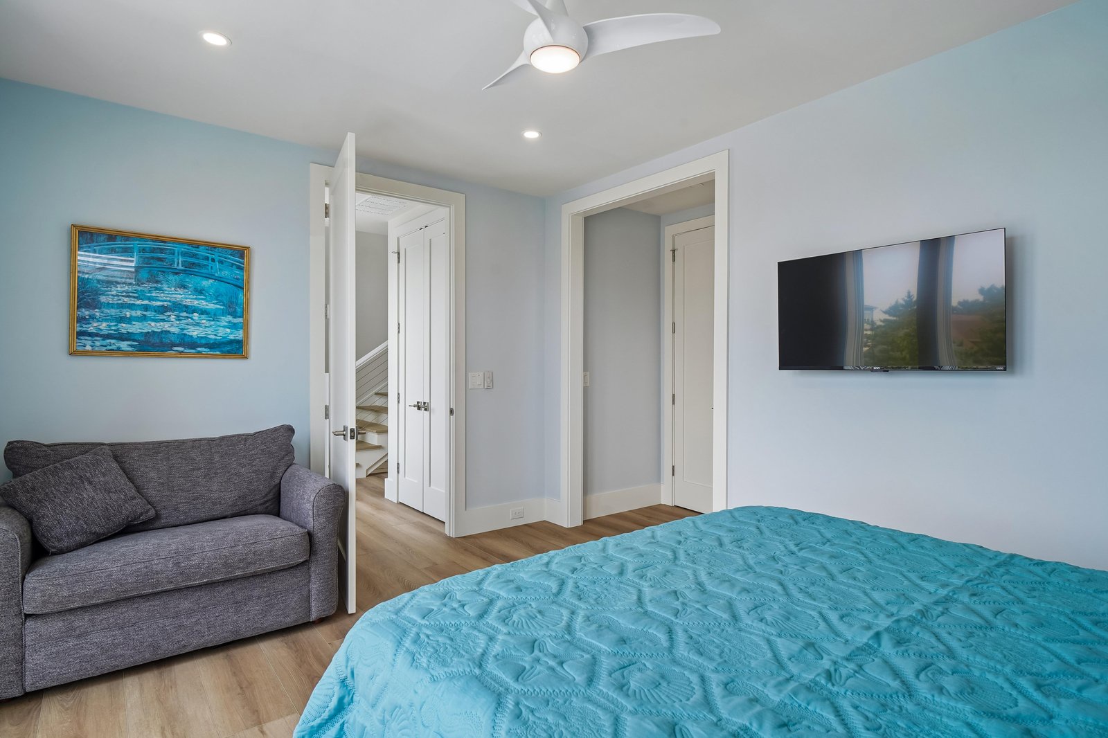 Relax in your coastal-inspired bedroom with airy blue tones, a cozy sofa for lounging, and modern touches like a ceiling fan and flat-screen TV. Soft natural light and thoughtful design create a serene escape.