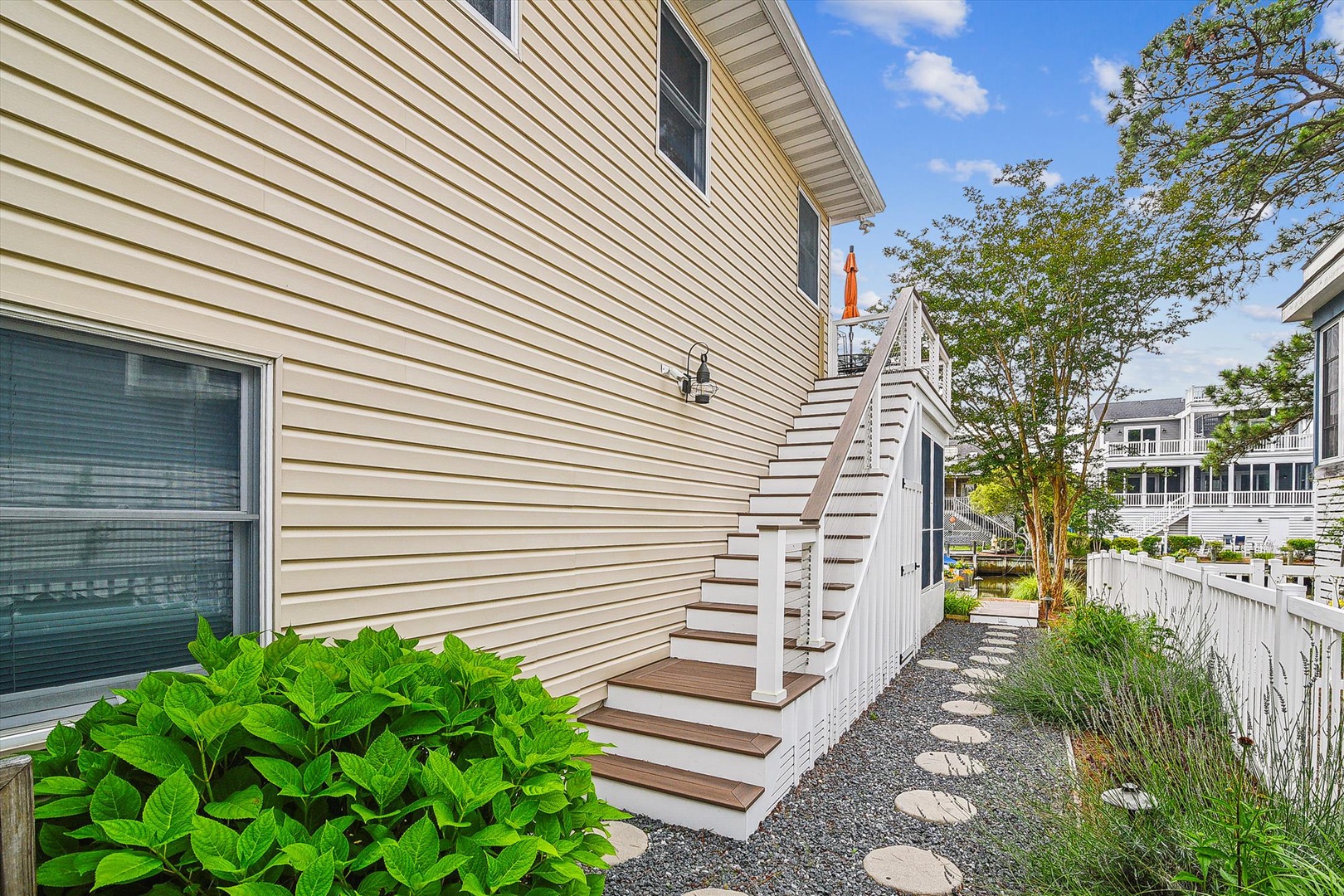 The property features a welcoming exterior entrance with a staircase leading to the home, nestled within a charming residential neighborhood surrounded by coastal greenery and mature trees.